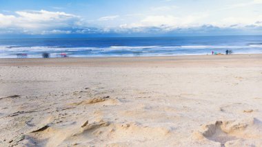 Sakin bir sahil sahnesi alçak gelgit sırasında kumlu bir sahili gösteriyor. Birkaç kişi sonbaharda bulutlu bir gökyüzünün altında uzun süre geziniyor..