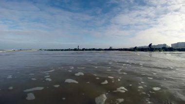 Kumlu bir sahil şeridinin sakin manzarası, kıyıya vuran sakin dalgalar, ve uzak arkaplanda görünen şehir binaları, modern kentsel yaşam ile birleşmiş doğanın güzelliğini somutlaştırıyor..