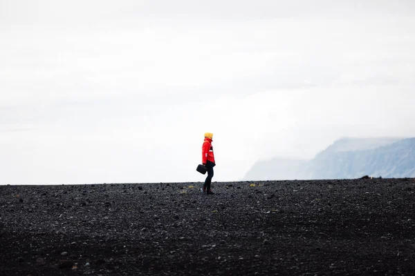 stock image A traveler in a red jacket standing on a black sand beach in Iceland. One person only, cold foggy Icelandic weather. 