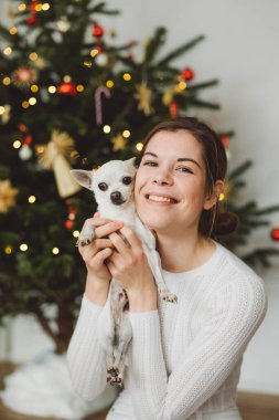 Beyaz giyinmiş, elinde küçük beyaz bir chihuahua ile Noel ağacının altında oturan gülümseyen beyaz kadın. Dikey fotoğraf.