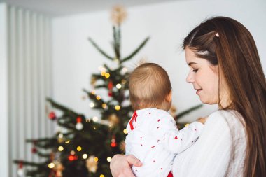 Genç anne ve kızının portresi, Noel 'de eğlenen küçük kız, Noel ağacını şenlikli kıyafetlerle süsleyen. Gülümseyen kız annesiyle oynuyor.. 