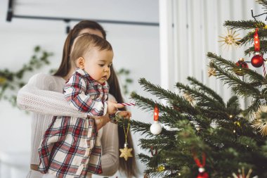 Genç anne ve kızının portresi, Noel 'de eğlenen küçük kız, Noel ağacını şenlikli kıyafetlerle süsleyen. Gülümseyen kız annesiyle oynuyor.. 