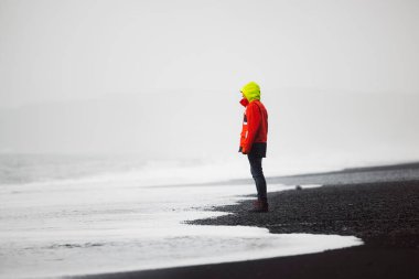 Su geçirmez ceketli ve pantolonlu adam siyah volkanik kumla sahilde duruyor, Reynisfjara, İzlanda 'nın güneyi. İzlanda doğası, Atlantik Okyanusu kıyısı, deniz kıyısı. Yüksek kalite fotoğraf
