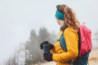 Belini kaldır kadın yürüyüşçü kameradan uzağa bakıyor elinde mavi bir termo şişeyle, sisli bir sonbahar gününde yürüyüş yapıyor., 
