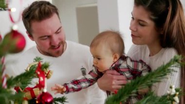 Beyaz giysiler içinde Noel ağacına bakan üç kişilik beyaz bir aile. Kırmızı bir elbise giyen bir bebek. Noel gününde sevgi dolu bir aile..