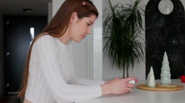 Gülümseyen beyaz bir kadın, mutfak tezgahında oturan bebek monitörüne bakıyor.. 