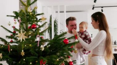 Beyaz giysiler içinde Noel ağacına bakan üç kişilik beyaz bir aile. Kırmızı bir elbise giyen bir bebek. Noel gününde sevgi dolu bir aile..