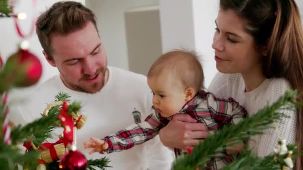 白い衣装を着たクリスマスツリーを見ている赤ちゃんの女の子と3人の白人の家族 お祝いの赤いドレスを着ている赤ちゃんの女の子 クリスマスの日に家族を愛する — ストック動画