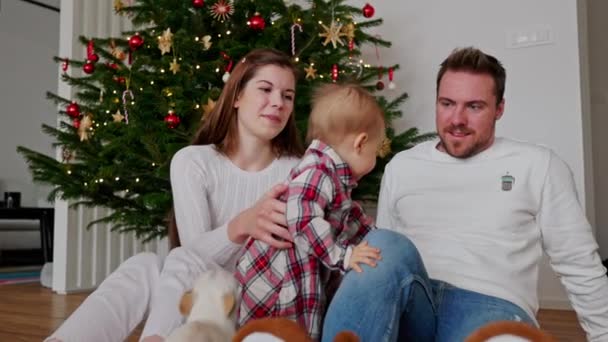 白い衣装を着たクリスマスツリーの下に座っている赤ちゃんの女の子と3人の白人の家族 赤ちゃんの女の子はお祝いの赤いドレスを着て クリスマスの日に家族を愛する — ストック動画