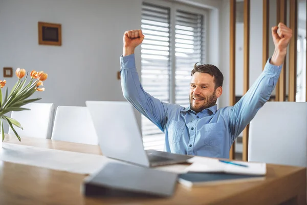 Excited caucasian man cheering for his team, watching a football game on his laptop, man celebrating a win with clenched fists.