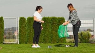 Caucasian women, mother and daughter picking up trash from the grass, one holding the green trash bag and other picking up trash with a stick. Cleaning the community.