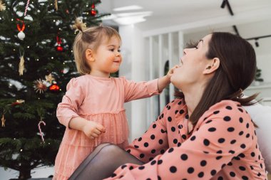 Young caucasian woman, mom, spending time with her toddler, young girl wearing a pink dress matching her mom. Mom and daughter decorating for Christmas together having fun. Mom holding a smiling