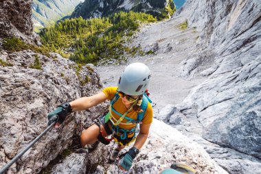 Caucasian woman mountaineer climbing up the mountain on via ferrata trail, high up in the mountain peaks. Woman wearing a protective head gear and uses a safety harness while she climbs.