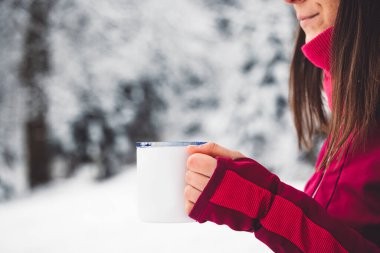 Beautiful young caucasian woman with brown hair, wearing a red jacket and a hat outside in the forest when its snowing. Woman walking around a snowy forest. Woman holding a to go cup with tea. 