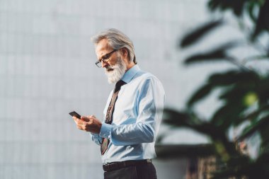 Senior business man with grey hair and a beard talking on the phone outside on a sunny day. CEO on the phone call outside.