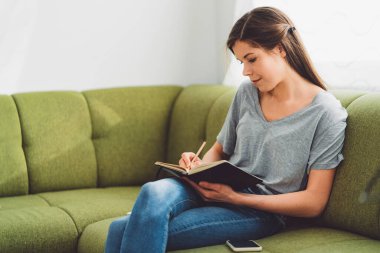 Young caucasian woman with long brown hair in jeans working from home, writing notes. Cheerful woman student studying at home. 