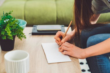 Young caucasian woman with long brown hair in jeans working from home, writing notes. Cheerful woman student studying at home. 
