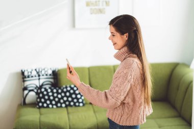 Young caucasian woman with long brown hair in pastel pink sweater and jeans working from home on her laptop and phone. Cheerful woman student studying at home. 