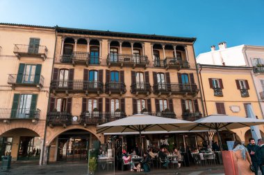Bir grup insan tenteli ve şemsiyeli bir binanın dışında oturuyorlar. Sahne hareketli ve hareketli, insanlar dışarıda eğleniyor.