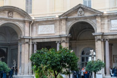 Bir grup insan, ortasında bir ağaç olan bir binanın önünde duruyor. Ağaç birkaç başka ağaç ve birkaç saksı bitkisiyle çevrilidir. İnsanlar birbirlerine yakın duruyorlar.