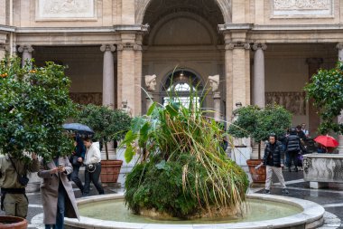 Bir grup insan avluda ortada bir çeşmeyle yürüyorlar. Çeşme bitkilerle çevrili ve içinde büyüyen büyük bir bitki var. İnsanlar şemsiye taşıyor.