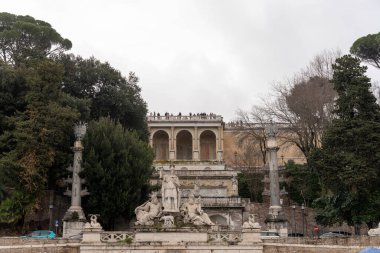 Önünde çeşme olan büyük bir bina. Çeşme ağaçlarla çevrili ve insanlar etrafında duruyor.