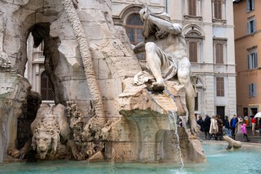 Arka planda çeşme olan bir kayanın üzerinde oturan bir adam heykeli. Heykel etrafta yürüyen ve ona bakan insanlarla çevrili. Sahne huzurlu ve sakin, heykel gibi.