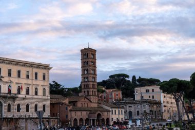 Arka planda uzun bir kule olan bir şehir caddesi. Gökyüzü bulutlu ve güneş batıyor.