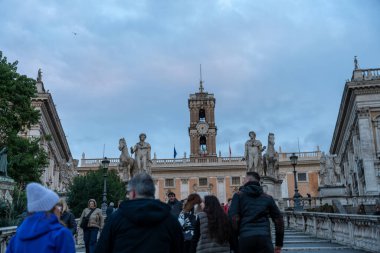 Bir grup insan büyük bir saat kulesinin önünde caddede yürüyorlar. Gökyüzü bulutlu ve atmosfer sakin ve huzurlu görünüyor.