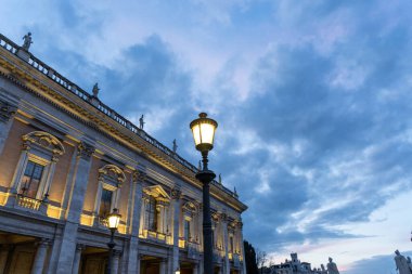 Önünde lamba direği olan büyük bir bina. Gökyüzü bulutlu ve lambadan gelen ışık binanın üzerinde parlıyor.
