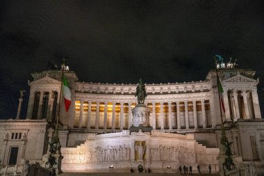 Üzerinde heykeller ve bayraklar olan büyük bir bina. Bina gece aydınlanıyor. Sahne muhteşem ve görkemli.