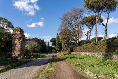 Yan tarafında taş bir duvar ve arka planında birkaç ağaç olan bir yol. Yol boş ve gökyüzü mavi.