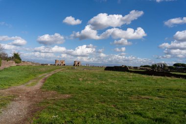 Toprak yolu ve arka planda birkaç bina olan bir arazi. Gökyüzü mavi ve bulutlu.
