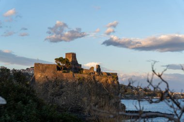Okyanusa bakan kayalık bir uçuruma tünemiş bir kale. Gökyüzü bulutlu, ama güneş hala bulutların arasında parlıyor, sıcak ve huzurlu bir atmosfer yaratıyor.