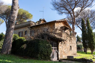 Ahşap kapılı ve küçük pencereli büyük taştan bir ev. Ev çalılar ve ağaçlarla çevrili.