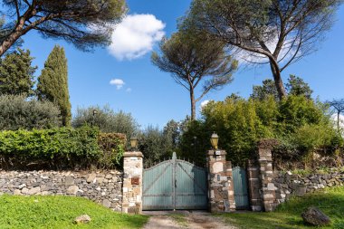 Üzerinde tabela olan bir kapı bir evin önündedir. Kapı mavi ve ev taş bir duvarla çevrili. Gökyüzü açık ve güneş parlıyor.