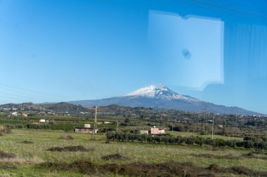Uzakta bir dağ sırası görülebilir, ön planda küçük bir kasaba vardır. Gökyüzü açık ve mavi, çimenler yeşil