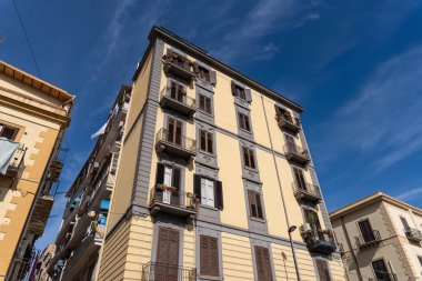 Balkonları ve pencereleri olan büyük sarı bir bina. Bina diğer binalarla çevrilidir. Gökyüzü mavi ve berrak