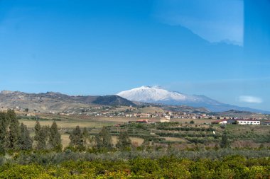 Tepesinde kar olan bir dağ sırası ve vadide küçük bir kasaba. Gökyüzü açık ve mavi