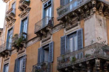 Mavi panjurlu ve balkonlu bir sıra kahverengi bina. Binalar eski ve kırsal bir havası var.