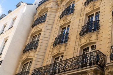 Balkonlu ve arkasında beyaz bir bina olan yüksek bir bina. Balkonda demir parmaklıklar var.