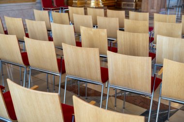 Büyük bir odada bir sıra tahta sandalye boş. Sandalyeler sıra sıra diziliyor, bazı sandalyeler öne bakıyor ve diğerleri uzağa bakıyor. Oda bir sınıf ya da konferans salonu gibi görünüyor.