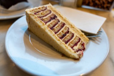 Sağ tarafında çatal olan beyaz bir tabakta bir dilim pasta. Pasta üçgen şeklinde kesilmiş ve çilek dolgulu.