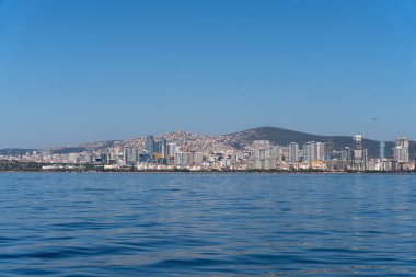 Bir şehrin ufuk çizgisi suya yansıyor. Gökyüzü açık ve su sakin. Şehir hareketlilikle dolup taşıyor ama su bir huzur ve sükunet hissi veriyor.
