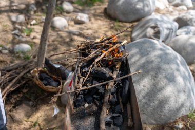 Ateş çukurunda bir odun ve kömür yığını yanıyor. Sahne açık havada ve arka plandaki kayalar doğal ortama ekleniyor.