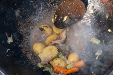 Bir tencere yiyecek kaşıkla karıştırılıyor. Yiyecek, havuç ve patates de dahil olmak üzere sebze ve et karışımı. Yiyeceklerin buharı yükseliyor, rahat ve davetkar bir atmosfer yaratıyor.