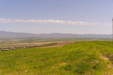 Uzaklarda birkaç evi olan geniş, açık bir arazi. Gökyüzü açık ve mavidir ve güneş ışıl ışıl parlar.