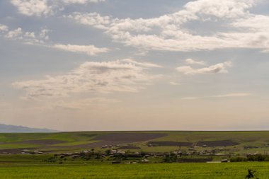 Uzaklarda birkaç evi olan geniş, açık bir arazi. Gökyüzü çoğunlukla açık ve birkaç bulut etrafa saçılmış durumda. Manzara, geniş bir araziyle huzurlu ve huzurlu.