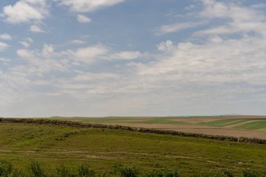 Mavi gökyüzü ve birkaç bulutu olan geniş, açık bir alan. Gökyüzü berrak ve parlak, ve çimenler yemyeşil. Sahne huzurlu ve sakin, insan aktivitesi belirtisi yok.