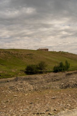 Evi ve ağaçları olan bir yamaç. Gökyüzü bulutlu ve güneş görünmüyor.